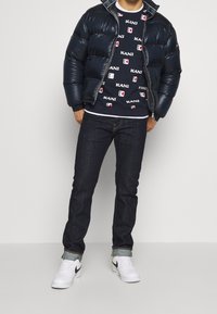 Giacca puffer blu navy, felpa con motivo e loghi, jeans scuri e sneakers bianche con dettagli neri. Materiale liscio e lucido sulla giacca.