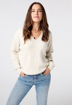 Crème geklede ribbelgebreide trui met een v-hals, lange mouwen, gecombineerd met lichtblauwe jeans. Opvallende gelaagde gouden kettingen en een armband.