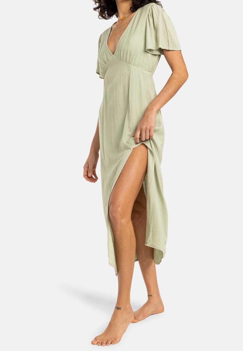 Femme portant une robe midi vert clair à col en V, manches volantées et fente haute à la cuisse, debout pieds nus sur fond blanc.