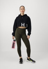 Felpa nera corta con scritte bianche, leggings verde oliva, sneakers nere con dettagli gialli, mentre tiene una bottiglia d'acqua rosa con logo nero.