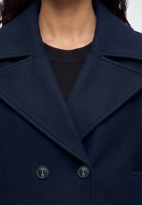 Femme portant un manteau bleu marine foncé à double boutonnage avec de larges revers sur un haut noir côtelé, visible du cou au milieu de la poitrine.