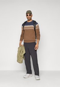 Homme portant un pull à motifs marron et bleu, un pantalon noir, des baskets blanches, des lunettes de soleil, une casquette et un sac à dos, tenant une veste olive.