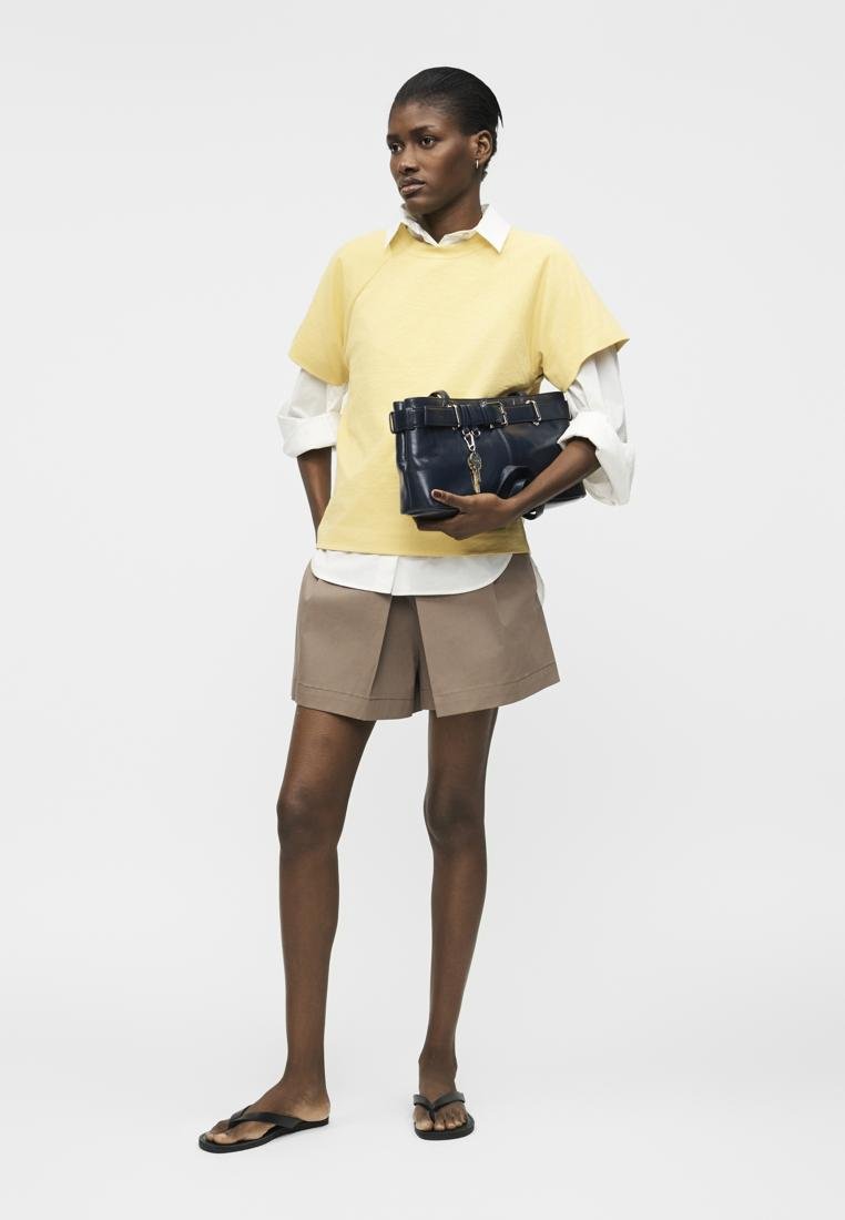 Femme portant un pull jaune à manches courtes sur une chemise blanche et un short beige, tenant un sac à main bleu marine, debout avec des tongs noires.