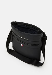 Black textured messenger bag with a zippered front pocket, adjustable strap, and a logo patch featuring red, white, and blue accents.
