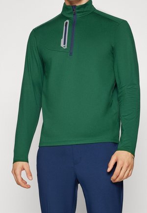 Homme portant un pull vert à manches longues avec une fermeture éclair quart de zip verticale sur la poitrine et un pantalon bleu marine.