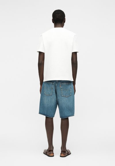 Άνδρας που στέκεται με την πλάτη προς την κάμερα, φοράει μια απλή λευκή κοντομάνικη μπλούζα, μπλε τζιν σορτς και καφέ σανδάλια σε λευκό φόντο.