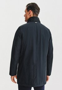 Granatowy płaszcz o wysokim kołnierzu, długich rękawach i prostej sylwetce. Gładka faktura z minimalnymi zdobieniami oraz szwem wzdłuż środka na plecach.