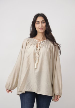 Kobieta o długich ciemnych włosach, ubrana w luźną beżową bluzkę z frędzlami oraz ciemne dżinsy, stojąca na tle jednolitego jasnoszarego tła.