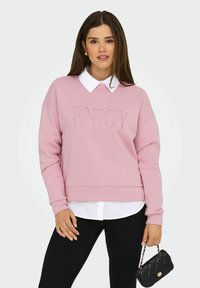 Vrouw in een roze sweatshirt met de tekst "ENJOY", een witte shirt eronder, zwarte broek, gouden oorbellen, ringen en een kleine zwarte gewatteerde handtas in de hand.