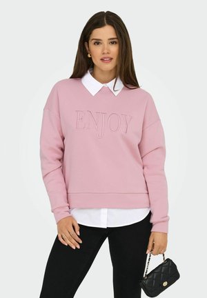 Donna che indossa un felpa rosa con la scritta "ENJOY", una camicia bianca sotto, pantaloni neri, orecchini d'oro, anelli e tiene in mano una piccola borsa trapuntata nera.