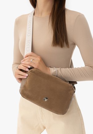 Mujer con top beige de manga larga y pantalones claros, sosteniendo un bolso cruzado de ante marrón con una correa blanca que lleva la etiqueta "RYŁKO".