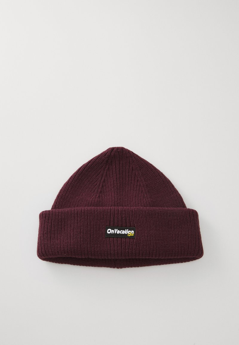 Gorro de malha bordeaux com textura canelada e uma dobra na parte inferior. Apresenta um retângulo preto com texto em amarelo e branco.