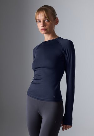 Haut à manches longues marine en tissu lisse et extensible avec une silhouette ajustée. Il possède des manches raglan et un col rond.