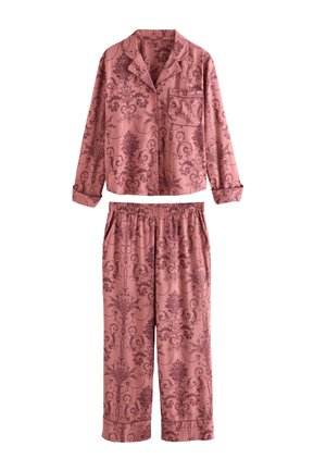 Woman smiling and standing barefoot wearing matching pink patterned long-sleeve pajama set in a softly lit room with flowers and furniture.