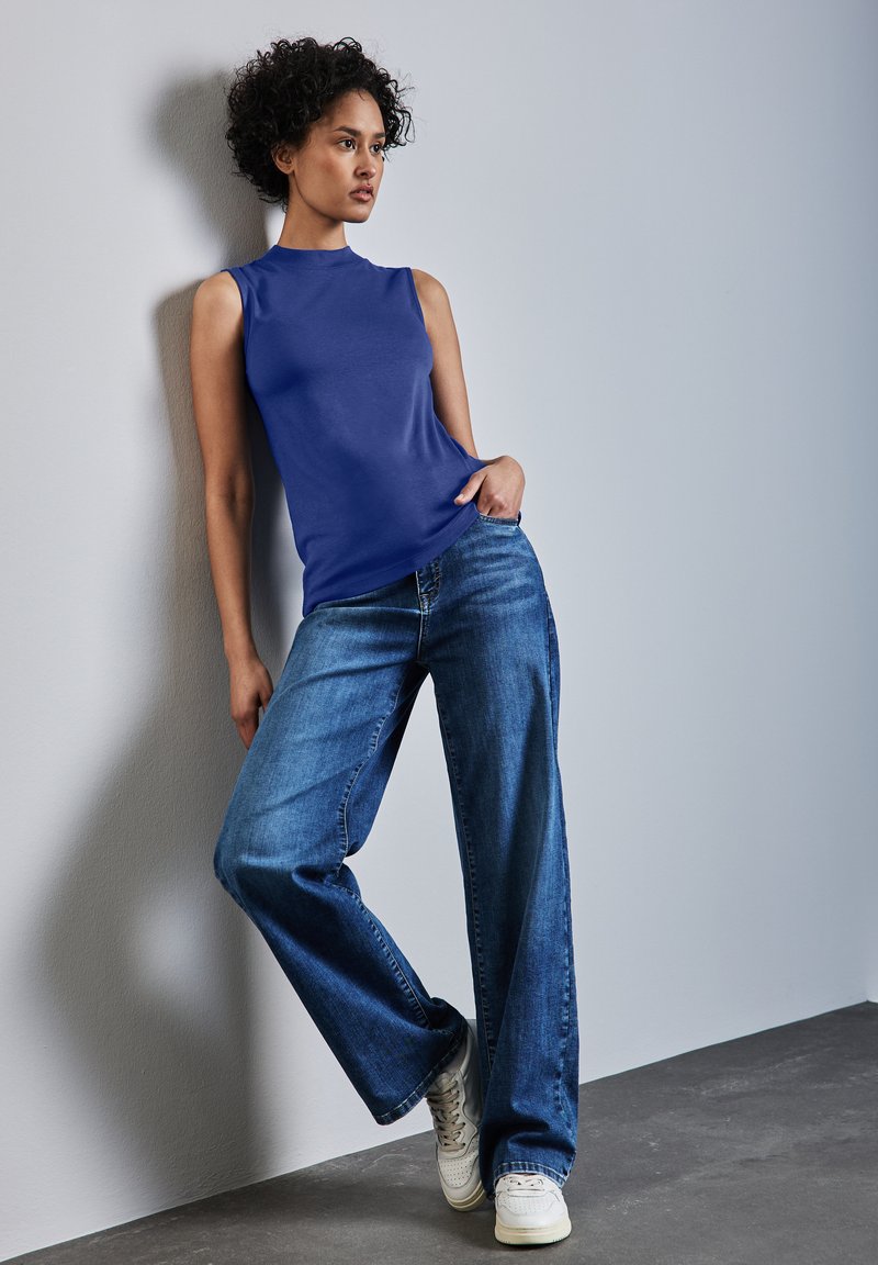 Ärmlös blå topp och vida blå jeans, med en mjuk textur och avslappnad passform; kombinerat med vita sneakers, mot en neutral bakgrund.
