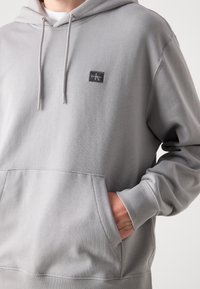 Grijze capuchontrui van zachte fleece, met een voorzak in kangoeroestijl, trekkoorddetails en een klein logo-patch op de borst.