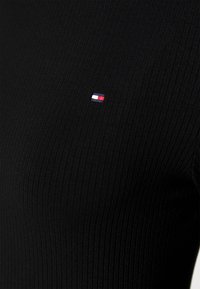 Svart ribbet topp laget av elastisk stoff, med en liten brodert logo i rød, hvit og blå på brystet.