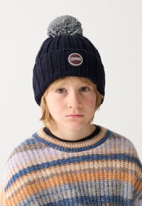 Beanie tricotat de culoare bleumarin, cu un pom-pom gri și logo-ul circular "Colmar" pe față, purtat cu un pulover striped multicolor.