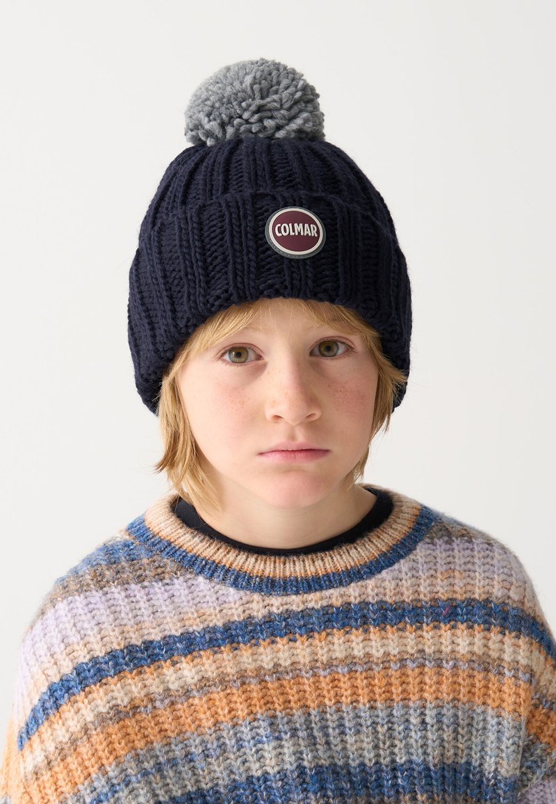 Beanie tricotat de culoare bleumarin, cu un pom-pom gri și logo-ul circular "Colmar" pe față, purtat cu un pulover striped multicolor.