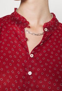 Blusa rossa con motivo floreale, colletto arricciato, chiusura frontale con bottoni e maniche corte. Presenta bottoni bianchi e una collana a catena argentata.