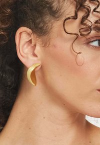 Zlaté, zakrivené náušnice s hladkým, kovovým povrchom. Navrhnuté v tvare polmesiaca, tesne obopínajúce ušnú lalôčik. Žiadne viditeľné prvky.