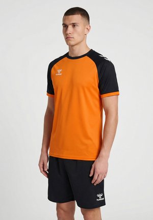 Jeune homme portant un t-shirt de sport orange et noir et un short noir avec des logos Hummel, debout devant un fond clair uni.