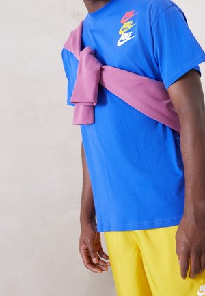 T-shirt bleu Nike avec des imprimés de logos multicolores, associé à un short jaune. Un pull rose est noué autour des épaules.