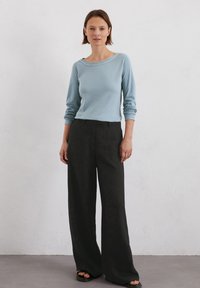 Helles blaues langärmliges Oberteil mit weitem Ausschnitt, kombiniert mit schwarzen weiten Hosen. Das Material wirkt glatt; der Schnitt ist lässig.