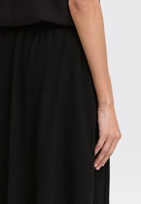 Jupe noire texturée avec une coupe loose. Présente une taille élastique et une main partiellement visible reposant sur le tissu.