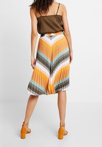 Falda midi plisada con un patrón en chevron en naranja, verde azulado y marrón, combinada con una camiseta sin mangas marrón y sandalias de tacón naranjas.