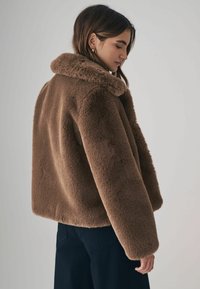 Brun faux fur jakke med bred krave, boxy form og lange ærmer. Teksturen er blød og fyldig, med en runding nederst.