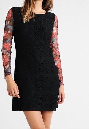 Femme portant une robe noire sans manches par-dessus un top transparent à manches longues avec des motifs floraux rouges, roses et bleus sur les manches.