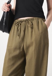 Olijfgroene linnen broek met een elastische tailleband en trekkoord, voorzien van zijzakken en een relaxte pasvorm. Zwarte handtas in één hand gehouden.