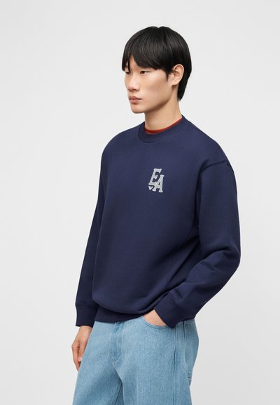 Νεαρός άνδρας που φοράει μπλε φούτερ με το λογότυπο "EA" στο στήθος και ανοιχτόχρωμο τζιν, στέκεται με το ένα χέρι στην τσέπη, κοιτάζοντας προς τα αριστερά.