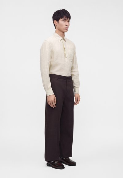Άνδρας στέκεται φορώντας κρεμ πουκάμισο μακρυμάνικο, σκούρο φαρδύ παντελόνι και σκούρα επίσημα παπούτσια σε απλό ανοιχτό φόντο.