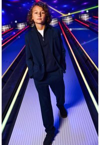 Marinblå kostym med en polotröja med dragkedja under, stående på en bowlingbana med färgglada ljus och en slät, vit yta.