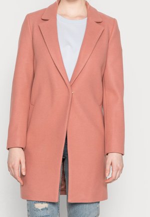 Personne portant un manteau rose poudré à un seul bouton par-dessus une chemise blanche et un jean bleu déchiré, debout devant un fond uni.