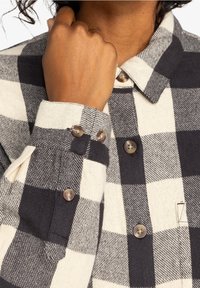 Ternet flannel skjorte med sort og hvidt mønster, der har knaplukninger og tekstureret stof. Synlige detaljerne på manchetter og krave.