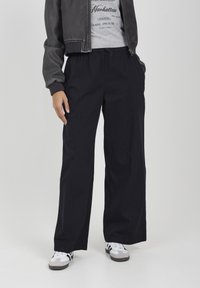 Persona che indossa pantaloni neri a gamba larga, maglietta grafica grigia, giacca scura e sneakers bianche, in piedi con le mani in tasca su uno sfondo bianco.