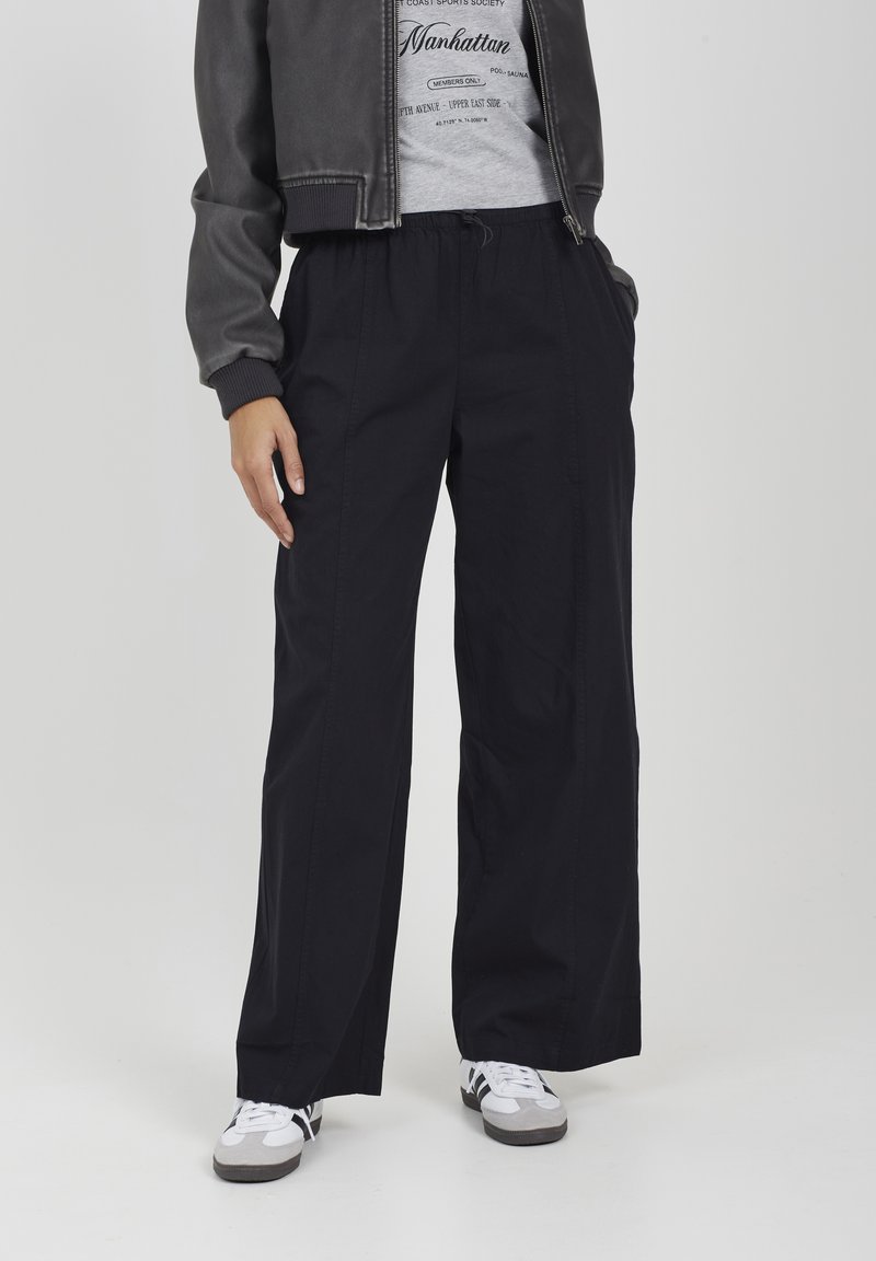 Persona che indossa pantaloni neri a gamba larga, maglietta grafica grigia, giacca scura e sneakers bianche, in piedi con le mani in tasca su uno sfondo bianco.