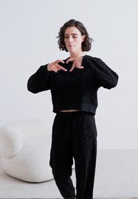 Svart cropped sweatshirt med teksturert blomstermønster, kombinert med matchende bukser med elastisk liv. Modellen står i en minimalistisk setting.