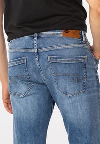 Mężczyzna w wyblakłych niebieskich dżinsach z przeszytymi tylnymi kieszeniami i czarnej koszuli, ręka spoczywa blisko prawego biodra.