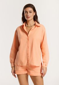 Camicia a maniche lunghe color pesca con chiusura a bottoni e dettagli ai polsini, abbinata a pantaloni corti coordinati, che mostrano una vestibilità rilassata e una consistenza morbida.