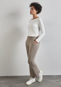 Maglietta bianca a maniche lunghe leggera e rigata, pantaloni beige slim-fit con tasche, indossati con sneakers bianche, su uno sfondo neutro.