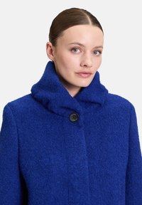 Manteau en laine texturée bleue avec un col haut, fermeture à un seul bouton noir, et une coupe légèrement oversize. Design minimaliste et tissu doux.