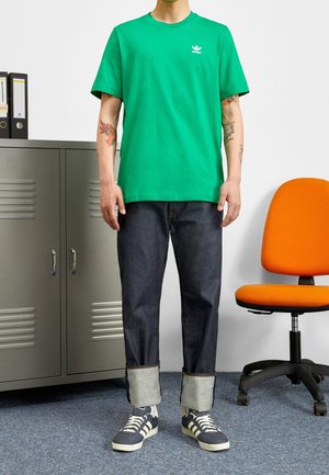 Grön bomullströja med en liten vit logotyp, matchad med mörk denimjeans som är uppvikt vid anklarna, och grå sneakers med vita detaljer.