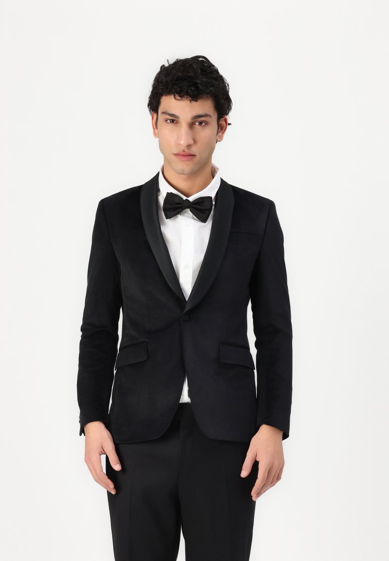 Jachetă de tuxedo neagră cu reveruri din satin, închidere cu un singur nasture și două buzunare frontale. Purtat�ă peste o cămașă albă și o cravată papion neagră.