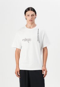 Hvid bomulds T-shirt med et gråt grafisk "nike" logo på forsiden og lodret tekst "International Sportswear" langs siden.