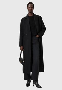 Abrigo largo negro de lana con un solo botón, combinado con un suéter negro, jeans de pierna ancha y botines de punta; sosteniendo un bolso negro.