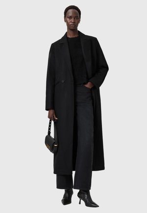 Svart långullrock med en enda knapp, ihop med en svart tröja, vida jeans och spetsiga ankellåga stövlar; håller en svart handväska.